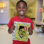 A young student holds up their art work and smiles for a photo in a school hallway